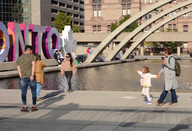 a young girl pulls her mother towards the toronto sign while she points at it, others are taking photos in front of the sign