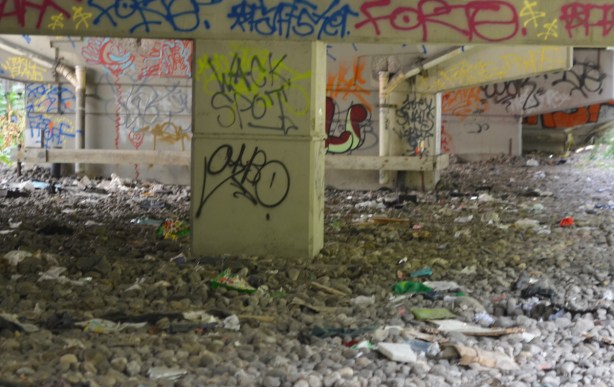 short pillars columns holding up a ramp to the Gardiner Expressway, street art and graffiti on them, rocks underneath, garbage in among the rocks 