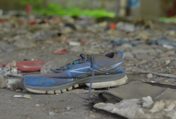 a black and blue running shoe with black laces on the ground under the Gardiner, among other pieces of garbage strewn about 