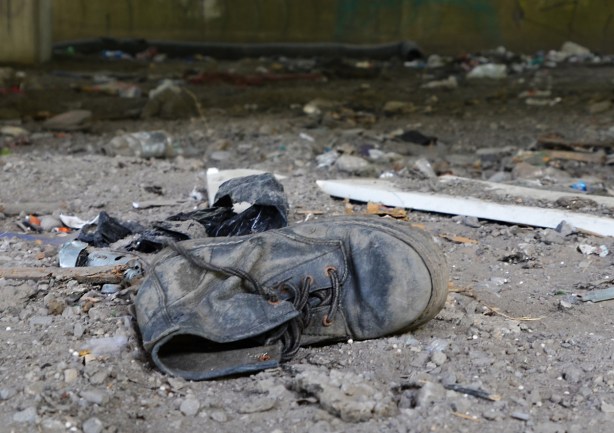 an old black boot lying on its side, laces undone, well worn, old, scuffed up, lying in the dirt, a black plastic garbage bag crumpled up is behind the shoe 
