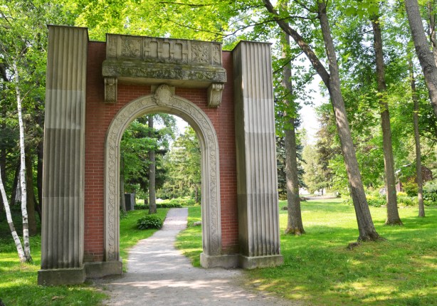 an arch entranceway of red brick and stone over a path through a garden with lots of trees and grass around it
