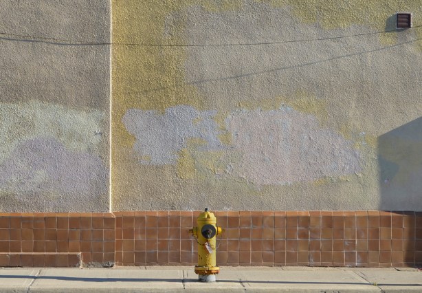 a yellow fire hydrant in front of a wall that is rust coloured tile on the bottom and grey stucco above. the stucco has been painted in splotches probably painting over graffiti 