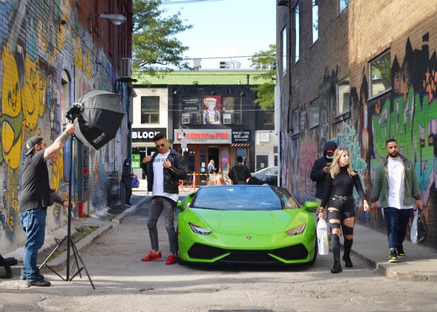 a young man in red shoes poses beside a green Ferrari parked in Graffiti Alley, being photographed, other people walking by including woman in ripped jeans 