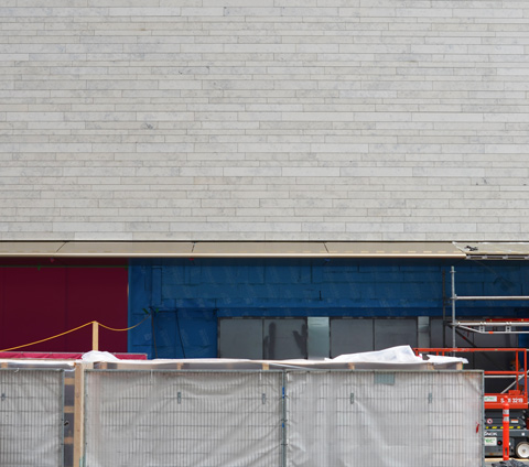 small section of wall in front of Holt Renfrew, grey cladding on top, part of pink and blue wall around the windows can be seen behind grey vinyl covered fence for construction 