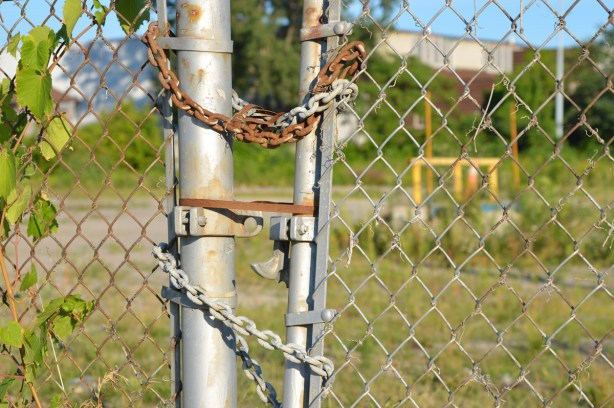 rusty chain holds a gate closed on chainlink fence, vacant lot behind