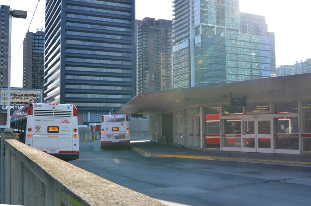 back of outside part of Shephard Subway station with a bus waiting by building, and an out of service bus parked towards the side, tall buildings of Yonge and Shephard area in the background 