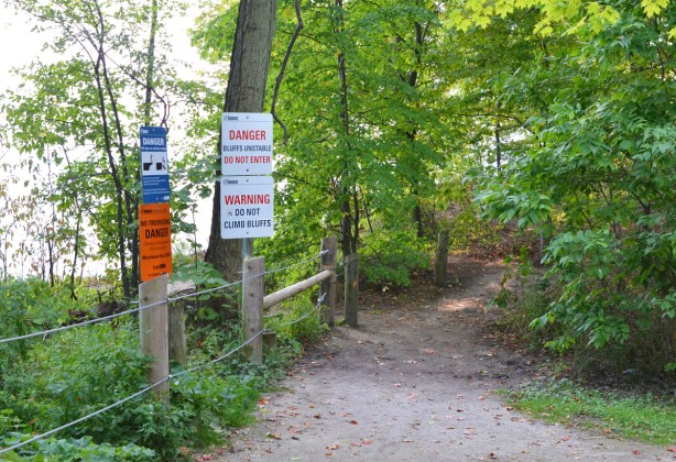 path through the woods with small fence on left. signs on left saying do not climb fence or cross over because of unstable ground, top of Scarborough bluffs, warning signs, 