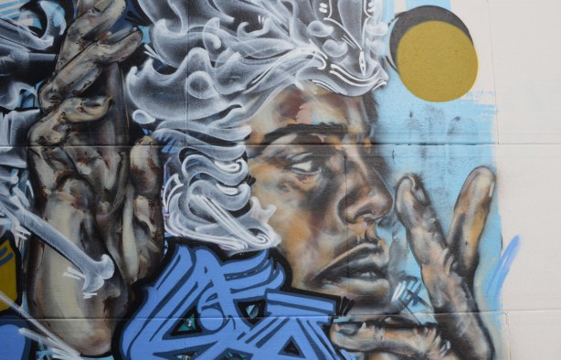 part of a mural of a black man with wavy white hair, blue text graffiti, and a yellow circle 
