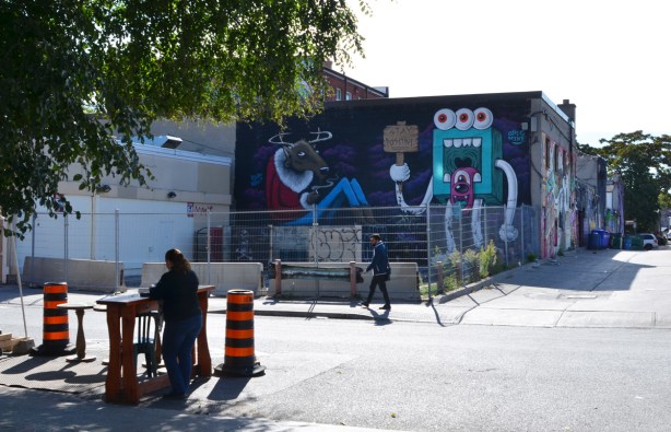 a woman stands by a table on the side of a road, across the street is a wall with two pictures on it, a cigarette smoking moose and a square thing with three eyes and long dangling white arms