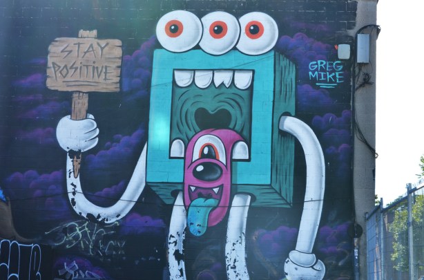 street art mural by Greg Mike of a turquoise square thing with a large square mouth and white teeth, three eyes across the top of its head, a pink droopy tongue with its own face and tongue, and long white arms and legs. In one hand it holds a sign that says Stay Positive 