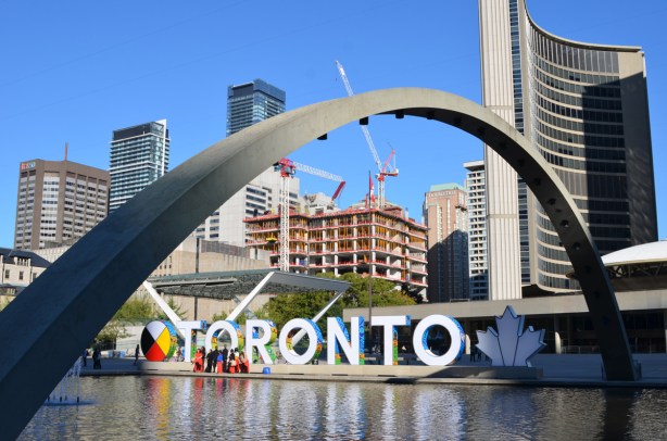 Nathan Phillips square with one arch prominent in the photo, reflecting pool, new Toronto sign, part of city hall, and construction of the new court house behind 