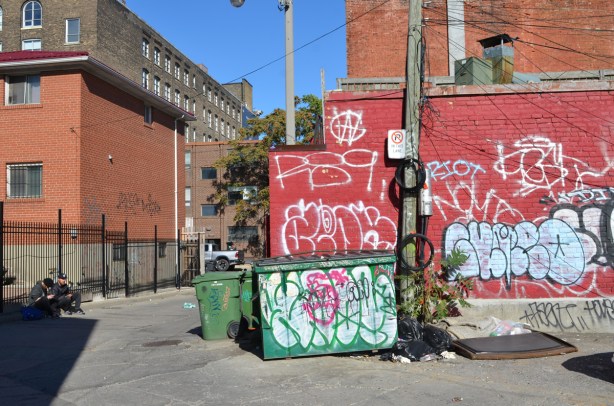 red brick wall with white tag graffiti, similar graffiti on green garbage bib two guys sitting on the curb on the other side of the lane 
