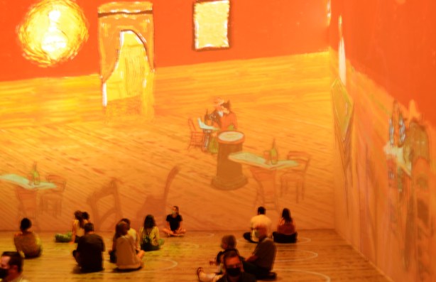 Vincent Van Gogh Immersive exhibit - people sitting on the floor with projections of images of inside of house with tables and chairs in orange and yellow tones