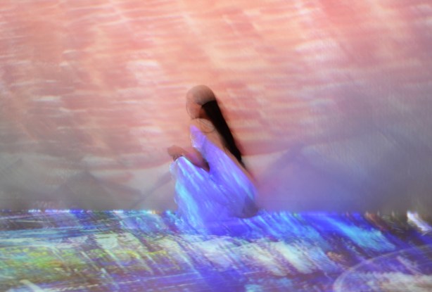 Vincent Van Gogh Immersive exhibit - a woman in a long white dress crouches beside the wall, images project onto her and her dress, pinks and purples 