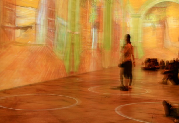 Vincent Van Gogh Immersive exhibit - a woman stands beside the wall, orange colours all around her including the floor, green images of windows and shutters too 