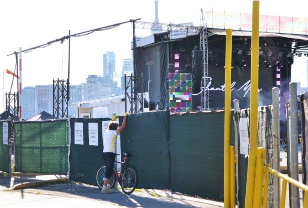 green covering on fence surrounding a temporary outdoor stage and theater. A man stands beside a bike, trying to look through gaps in the fence 