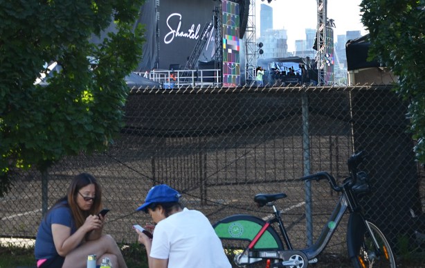 two people sitting on the ground looking at their phones in the foreground, fence between them and a singer rehearsing on a stage behind them 