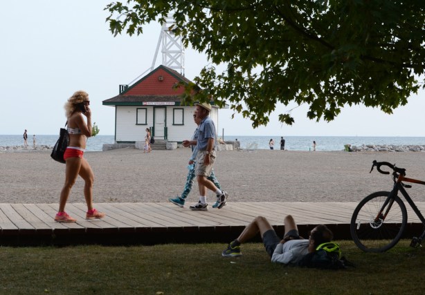 a man lies on the grass and watches people passing by on the boardwalk including a woman in a bathing suit and red shoes while talking on her phone, in front of Leuty Lifeguard station 