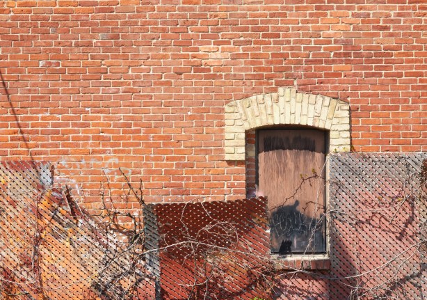 red brick wall, boarded up window with curved upper window frame, 4 wood trellises in front in different shades of rust and brown
