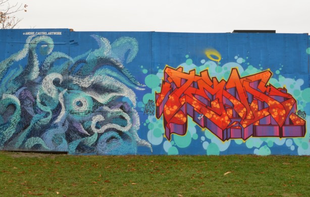 two murals side by side on blue hoardings, on the left is a blue monster by andre castro and on the right is an orange and red text graffiti 
