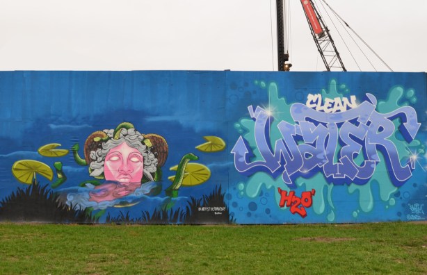two murals on hoardings around new water treatment plant by Ashbridges, on the left a very pink faced lady with grey curly hair in the water with little green dragons and lily pads. on the right, text painting that says clean water H 2 O