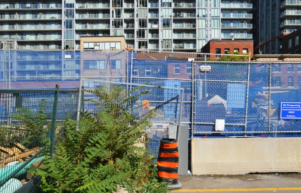 blue construction fence around a hole at a work site, row of storefronts across the street in the next level and a tall apartment building behind that 