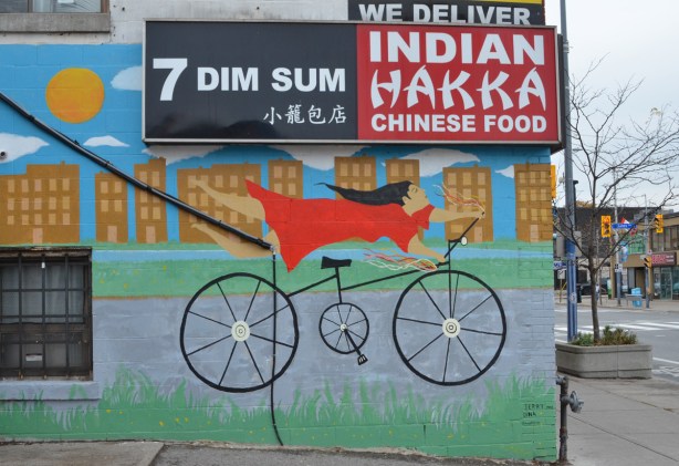 on the side of a Chinese restaurant, a mural of a woman on a bike. She's holding the handle bars but her legs are straight out behind her. She's wearing a red dress and has long black hair. There are three signs on the restaurant, First, Indian Hakka Chinese Food, second, 7 dim sum, and third, we deliver 
