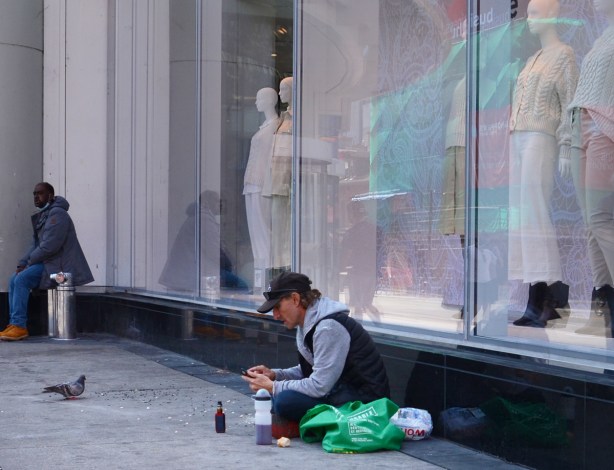 windows of H & M store at yonge and Dundas, with femaile mannequins, sitting on the sidewalk in front of them is a man feeding pigeons, another man sits nearby 