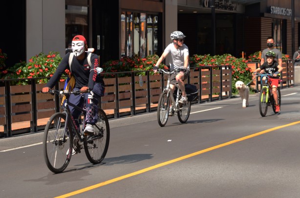 three people on bikes on Yonge street during streets open, the man in front is wearing an anonymous mask 