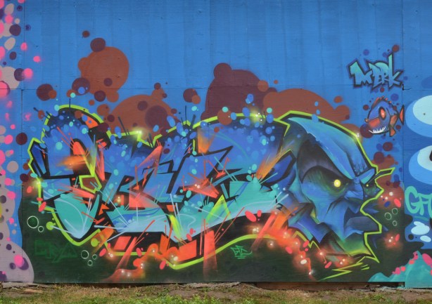 mural by Cruz 1 on hoardings around a construction site near Ashbridges, a blue face and lots of colours around word that says Cruz, plus a tiny fish in top right