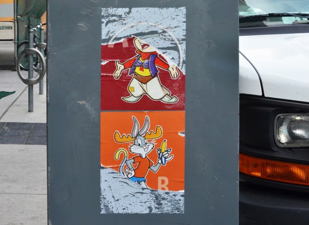 two coloured posters on a box on the street, both torn. On top, on red background is bugs bunny. on bottom on orange background is bugs bunny eating a banana