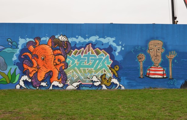 two murals on hoardings around new water treatment plant by Ashbridges, an orange octopus and a man in a red and white striped T shirt with words rise up 