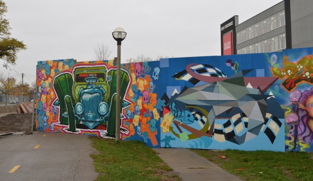 wallnoize street art murals on blue hoardings around new water treatment plant, one end of the hoardings, by the bike trail, an old green car by spud, and a graphic in neutral tones, 