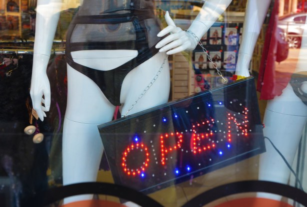 open sign in red and blue lights in the window of an adult store, beside white mannequin with very small black bikini bottoms and mesh top 