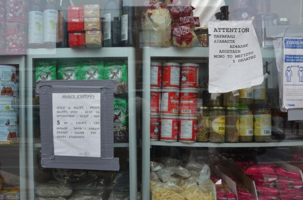 the window of a Greek grocery store, with food, also signs re covid rules in Greek. for sale, oregano, Nescafe coffee, tomato paste, 