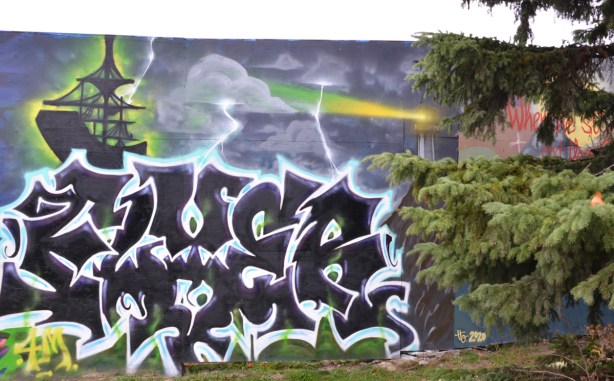 black and white wildstyle mural on blue hoardings, pine tree beside