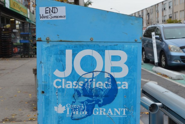 large dark blue stencil of a skull in profile on a blue newspaper box. There is also a sticker that says end white supremacy