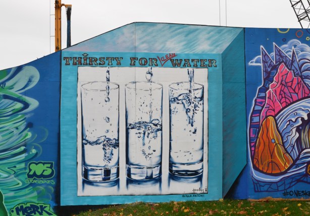 three tall glasses of water, each with a different amount, with the words thirsty for clean water, in a turquoise box, a mural 