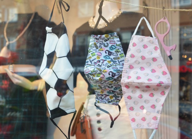 three masks on display in a store window.  One has a soccer ball pattern, another is pink with little strawberries.  The one in the middle is covered with a jumble of letters of the alphabet 