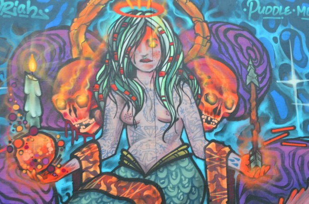 mural, long haired woman with nose ring, topless, upper body covered with tattoos, with scarves over her arms, halo on head, skulls beside her and a burning candle in each hand 