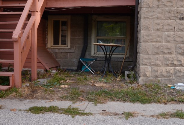 a folding chair and a small round table set up under a back porch behind a stone building, dark