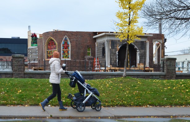 a woman pushing a stroller and holding a cup of coffee, walks past the shell of a ground floor of a brick church with a brick and stone porticao front entrance, fake glass for stained glass window, workmen still constructing it, a film set, being built right beside the sidewalk