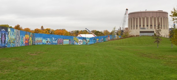 round Ashbridges water treatment tower with lots of green grass in front, a blue hoardings wall around the construction of new parts of water treatment plant, with lots of street art murals painted on the hoardings