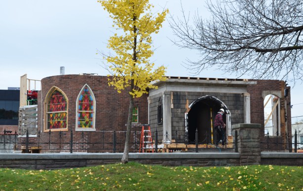 the shell of a ground floor of a brick church with a brick and stone porticao front entrance, fake glass for stained glass window, workmen still constructing it, a film set, 