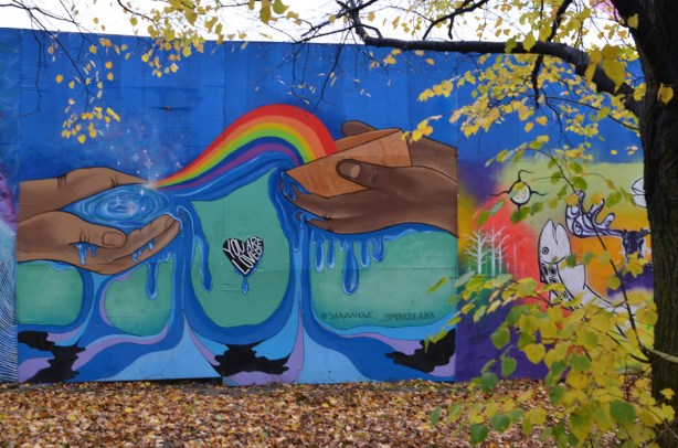 mural on the hoardings around the ashbridge bay water treatment facility expansion construction site. the words you are loved are in a heart shaped space in the middle, brown hands holding a shallow brown bowl fromwhich a rainbow is pouring out into the smaller cupped hands of another person 
