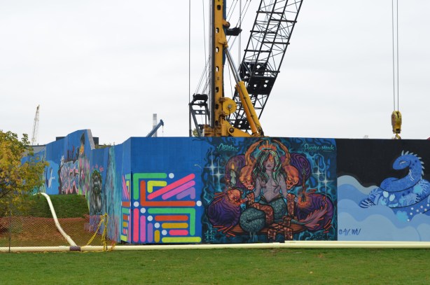 wallnoize street art murals on blue hoardings around new water treatment plant, cranes behind the fence, murals at the corner