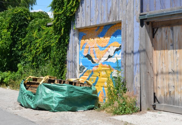 large green plastic bag on the ground filled with wood palletts, in an alley, in front of a garage door with street art on it 