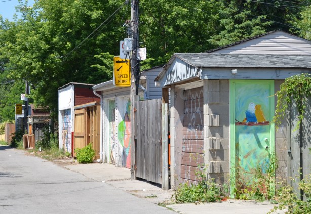 garages in an alley with garage doors that have been painted with street art, the garage in front has a light greens side door with two birds sitting on a braanch painted on it 