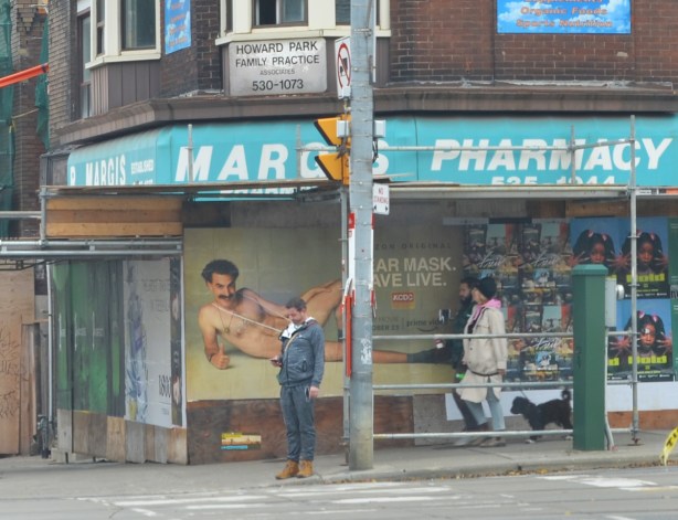 a man stands at an intersection waiting for traffic lights to change, he's in front of Borat poster, wear a mask, save live, man sands where he covers the mask 