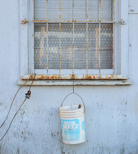 a window with on old rusty metal grille covering it, and a white bucket hanging by a chain 
